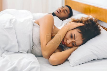 Black woman seeking peace by covering her head with arms while lying in a cozy bed next to her snoring partner, indicating stress or headache