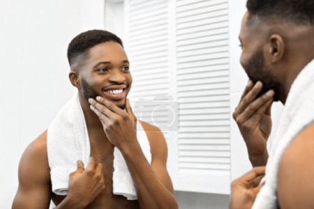 Foto de Hombre afroamericano con una toalla cubierta sobre sus hombros está acariciando su cara seca, lo que indica una rutina de cuidado de la piel o aseo - Imagen libre de derechos