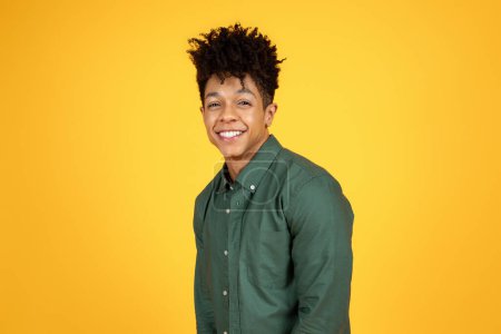 Téléchargez les photos : Joyeux jeune homme afro-américain rayonnant avec un large sourire engageant, debout devant une toile de fond jaune frappante - en image libre de droit