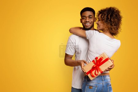 Téléchargez les photos : Gai noir guy eu cadeau de son amour copine, tenant boîte cadeau et câlins avec femme, fond orange - en image libre de droit