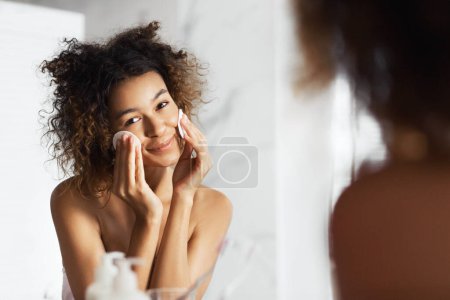 Téléchargez les photos : Belle fille afro-américaine nettoyant visage avec des tampons de coton regardant dans le miroir à la salle de bain. Tous les types de peau tonisation concept - en image libre de droit