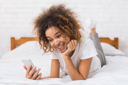 Una joven afroamericana de pelo rizado mira atentamente su teléfono móvil mientras se apoya en la cama con una expresión contemplativa