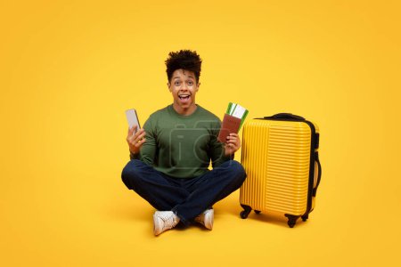 Foto de Un joven afroamericano se sienta en el suelo con su teléfono en una mano y pasaporte con tarjetas de embarque en la otra, junto a una maleta de color amarillo brillante, expresando emoción, alegría por viajar - Imagen libre de derechos