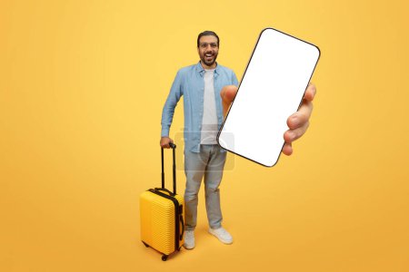 Foto de Hombre indio excitado sosteniendo una maleta y presentando un teléfono inteligente con pantalla en blanco para publicidad - Imagen libre de derechos
