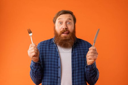 Photo for Hungry redhaired bearded guy holding knife and fork looking at camera and licking lips on orange studio background. Portrait of funny man ready for tasty dinner, having food cravings and temptation - Royalty Free Image