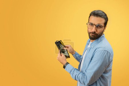 Photo for A worried indian man in a denim shirt displays an empty wallet, emphasizing financial concepts on a yellow background - Royalty Free Image