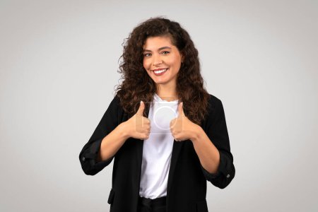 Téléchargez les photos : Image lumineuse et positive d'une femme souriante aux cheveux bouclés dans un blazer donnant les pouces vers le haut - en image libre de droit