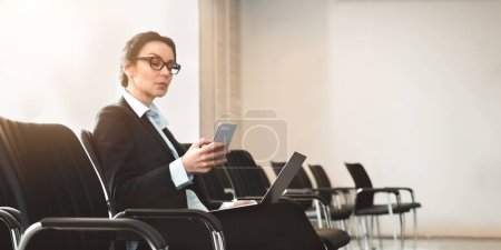 An elegant businesswoman uses her smartphone and laptop simultaneously in an empty conference room, exemplifying efficiency
