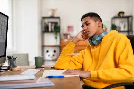 Photo for Weary black teenager guy in yellow hoodie and headphones takes moment to rest his eyes while sitting at work desk with computer and notebook - Royalty Free Image