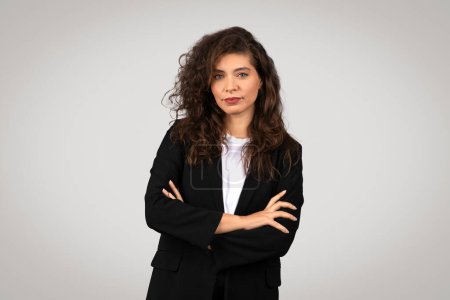A professional curly-haired woman in business attire with a confident posture and a neutral background