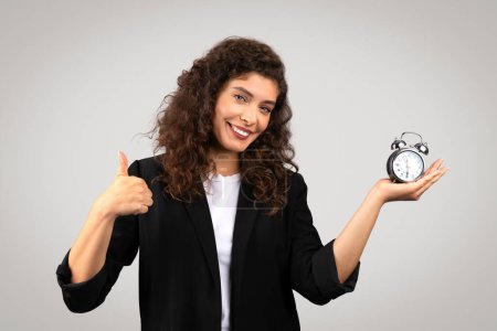 Mujer de negocios sonriente sosteniendo un reloj despertador y dando un pulgar hacia arriba, simbolizando la gestión del tiempo