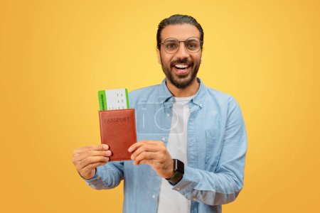 Téléchargez les photos : Homme indien souriant présentant un passeport avec des cartes d'embarquement sur fond jaune, suggérant la préparation au voyage - en image libre de droit