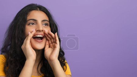 Excited woman in a yellow t-shirt shouts with her hands cupped around her mouth, purple background
