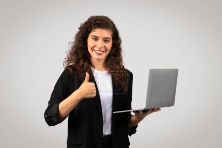 Téléchargez les photos : Une femme confiante aux cheveux bouclés, tenant un ordinateur portable et donnant un pouce vers le haut, isolée sur un fond gris - en image libre de droit