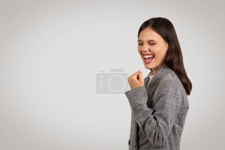 Téléchargez les photos : Délicieuse jeune femme en veste de poule classique, poing pompé dans la célébration, riant joyeusement, isolée sur fond gris clair, espace de copie - en image libre de droit
