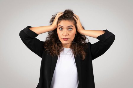 Photo for A young woman in a black blazer looks stressed, holding her head with both hands, against a neutral background - Royalty Free Image