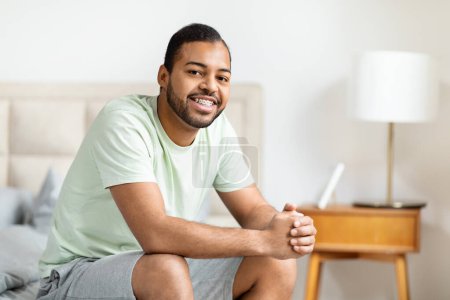 Ein fröhlicher afrikanisch-amerikanischer Mann sitzt auf der Bettkante und hält ein digitales Tablet mit einem warmen, einladenden Lächeln