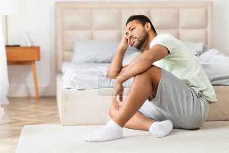 Una imagen que captura a un hombre negro deprimido con atuendo casual sentado con las piernas cruzadas en la alfombra junto a una cama en un ambiente acogedor dormitorio