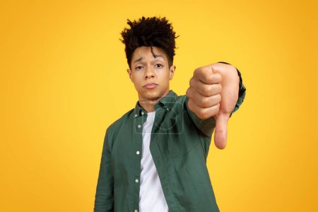 Joven afroamericano disgustado mostrando el gesto de los pulgares hacia abajo con una mirada escéptica sobre un fondo amarillo