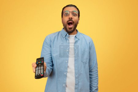 A surprised indian man holding a remote control and expressing shock on a yellow background