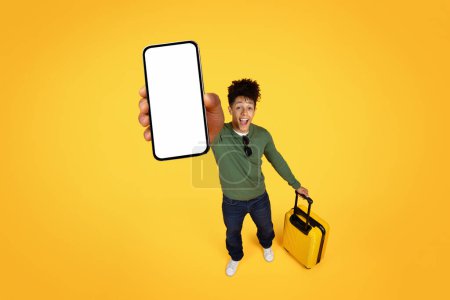 Cheerful young black man displaying a blank phone screen while holding a yellow suitcase, isolated on yellow