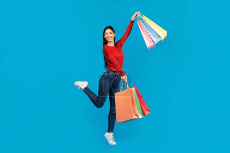Foto de Mujer joven emocionada saltando alegremente en el aire mientras sostiene bolsas de compras, mostrando su entusiasmo por sus compras, posando sobre fondo azul - Imagen libre de derechos