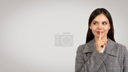 Foto de Mujer de negocios pensativa en blazer a cuadros grises con el dedo en los labios, mirando hacia el espacio libre pensativamente, sugiriendo silencio o contemplación, sobre fondo claro - Imagen libre de derechos