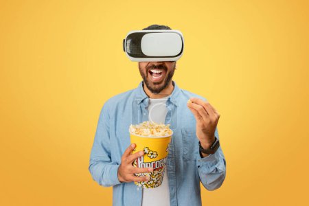 Eastern man engrossed in virtual reality, enjoying popcorn while immersed in a simulated world against a yellow backdrop