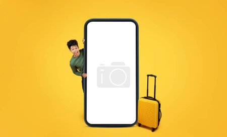 A african american guy playfully peeking from behind an oversized smartphone with a suitcase on yellow backdrop