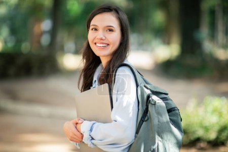 Photo for Confident smiling young european woman with a beaming smile, clutching a laptop, wearing a light blue jacket, and shouldering a backpack, walking in a sunny park, outside - Royalty Free Image