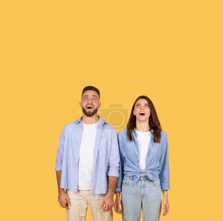 Foto de Un hombre y una mujer con expresiones de asombro y sorpresa, mirando hacia arriba sobre un fondo amarillo que amplifica sus emociones - Imagen libre de derechos