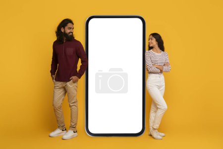 Téléchargez les photos : Homme et femme de chaque côté d'un grand écran de smartphone vierge, simulant l'interaction sur un fond jaune - en image libre de droit