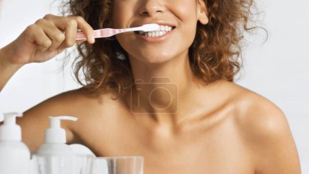 Téléchargez les photos : Jeune femme afro-américaine souriante avec brosse à dents nettoyant les dents dans la salle de bain. Concept de blanchiment des dents - en image libre de droit