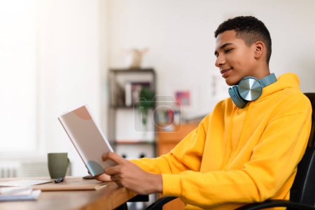 Photo for A relaxed young man in a yellow hoodie reads a book with headphones around his neck at a home office desk - Royalty Free Image