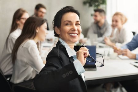 Foto de Una mujer confiada con gafas en la mano sonríe a la cámara con una reunión de equipo en segundo plano, sugiriendo liderazgo y un ambiente de trabajo positivo - Imagen libre de derechos