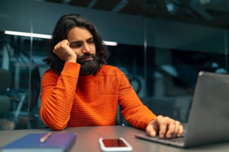 Hombre de mediana edad en un suéter naranja parece aburrido mientras usa una computadora en un escritorio