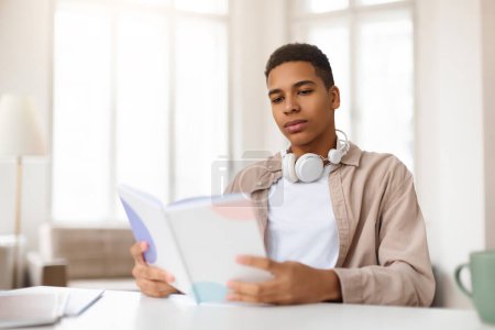 Foto de Hombre joven y seguro con auriculares alrededor de su cuello lee un libro mientras está sentado en un escritorio de madera con papelería y una taza de café - Imagen libre de derechos