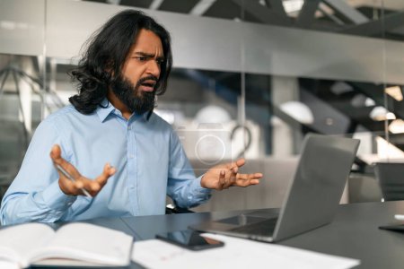Un homme aux cheveux longs et une chemise bleue montrant des signes de frustration et de confusion en regardant un écran d'ordinateur portable