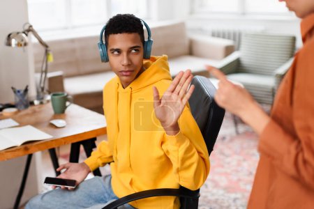 A young man wearing a yellow hoodie and headphones makes a stop gesture towards an unseen person while at his desk