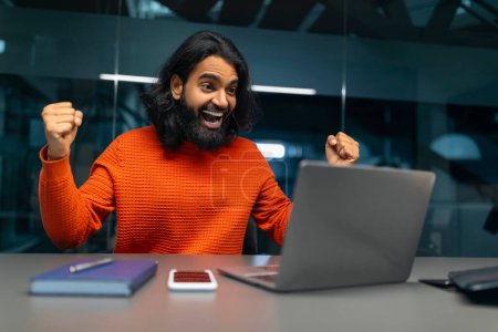 Téléchargez les photos : Homme enthousiaste avec les poings levés dans la joie devant son ordinateur portable à un bureau - en image libre de droit
