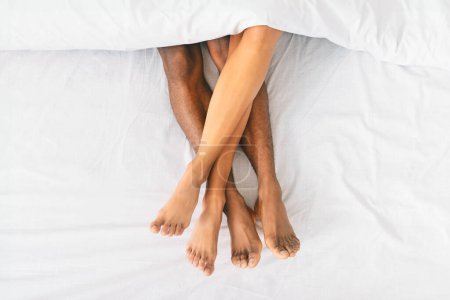 Téléchargez les photos : Couples afro-américains jambes entrelacées sous un drap de lit blanc croustillant, symbolisant l'intimité et l'espace partagé dans une relation - en image libre de droit