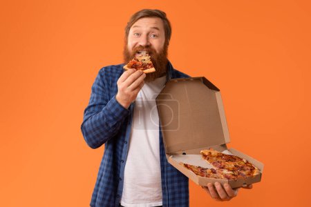 Photo for Fast food eater. Cheerful redhaired bearded guy bites into a slice of pizza in studio with orange background, while holding delivery box and smiling to camera. Concept of junk food pleasure - Royalty Free Image