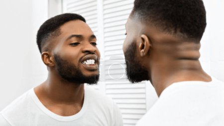 African american guy is brushing his teeth attentively while observing himself in a bathroom mirror, indicating hygiene
