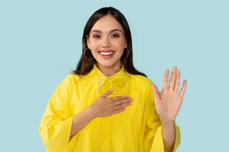 Happy glad sincere young woman work, raises hand, swearing an oath. Expression embodies commitment and trustworthiness, ideal for campaigns, ads, and assurance-themed visuals, blue background.