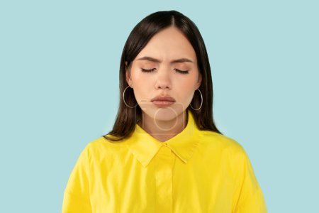 Upset unhappy emotional young woman, with eyes closed showing a vulnerable moment of sadness, dramatically grimacing against blue studio background