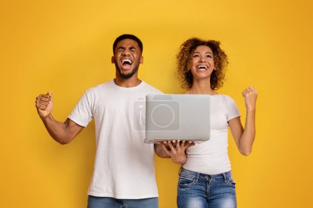 Foto de Un hombre y una mujer afroamericanos extáticos celebrando el éxito con una computadora portátil sobre un fondo amarillo - Imagen libre de derechos