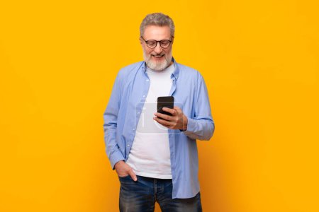 Téléchargez les photos : Homme âgé européen avec les cheveux gris défilant à travers les messages sur le téléphone portable, debout vérifier les e-mails et les applications dans le contexte de studio jaune. Médias sociaux et communication - en image libre de droit