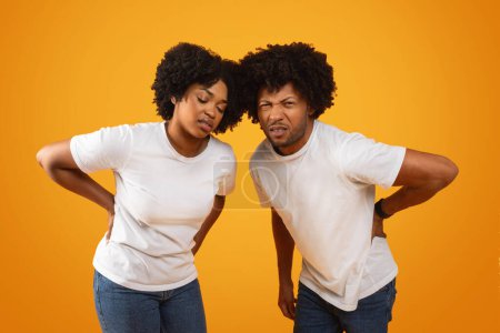 Foto de Desdichados millennials afroamericanos cónyuges sufren de dolor de espalda baja. Hombre y mujer negros cansados y enfermos que tocan su espalda, fondo amarillo. Mala postura, osteoporosis, tensión muscular - Imagen libre de derechos