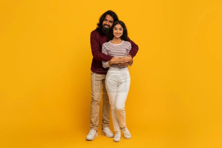 A smiling couple stands close together, hugging in front of a vibrant yellow backdrop, portraying warmth and affection