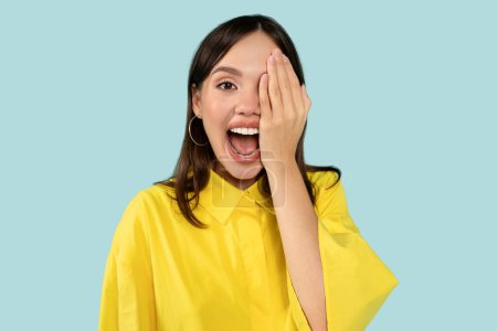 Photo for Portrait of happy surprised woman covering one eye with palm, smiling broadly expresses positive astonishment, laughs indoors against blue studio background - Royalty Free Image